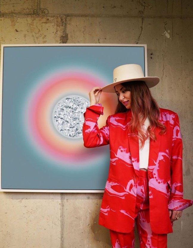 Lauren Baker stands in front of her artwork holding the brim of her hat and wearing a brightly-coloured suit