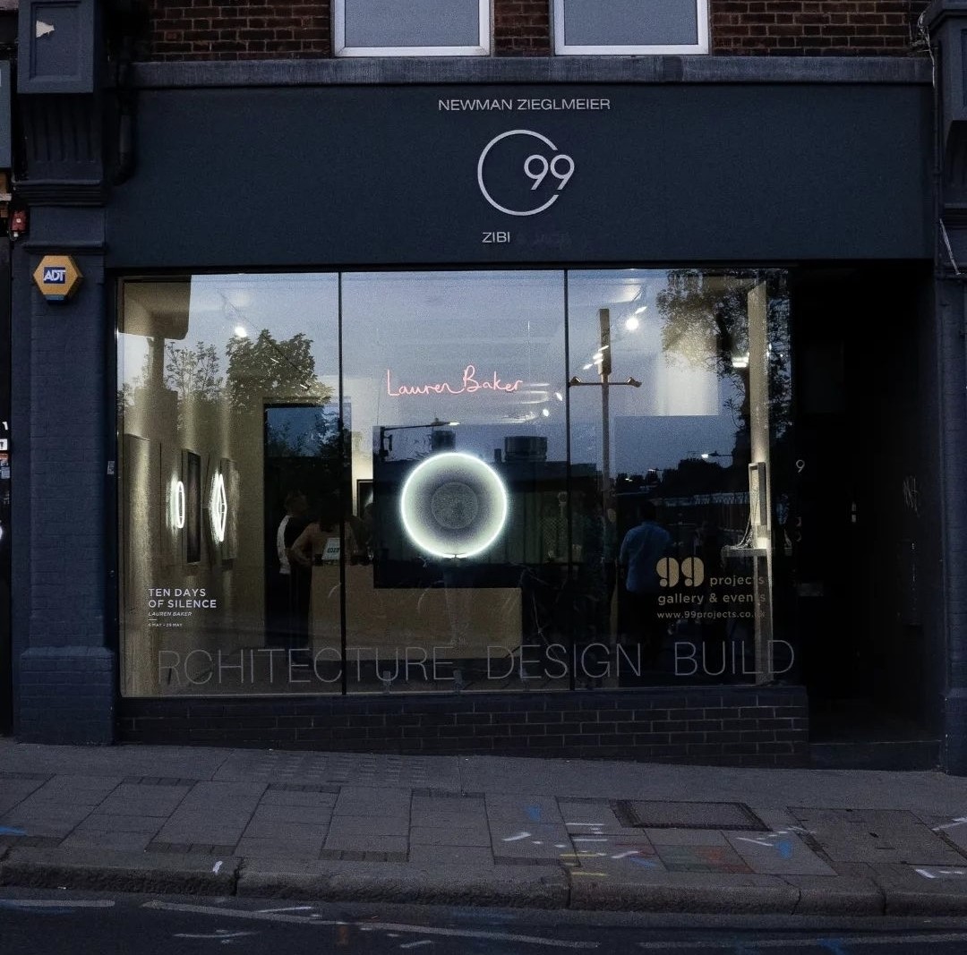 An external shot of the 99 Projects gallery. In the window there is Lauren Baker's name in pink neon lettering and a separate circular neon painting